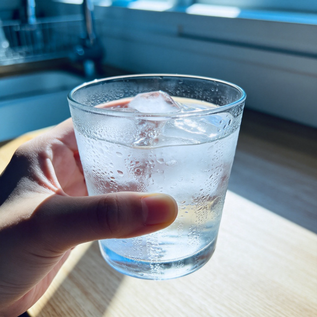 Close-up shot of a person's hand holding a clear glass filled with water and ice cubes, with condensation droplets on the outside of the glass. The background is a sunlit kitchen counter. The focus is on the refreshing, cold look of the drink. No text.