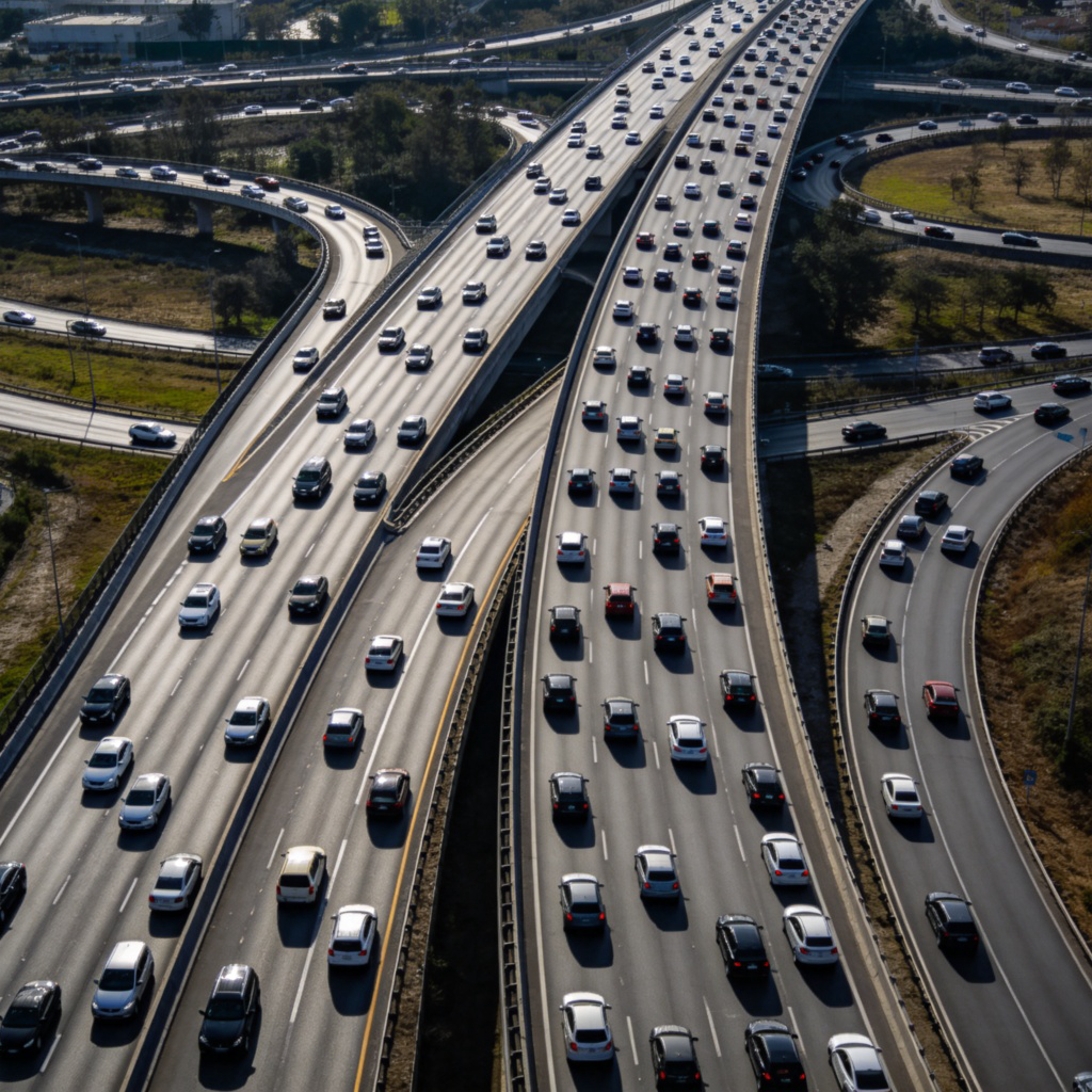 A wide-angle top-down view of a large, busy highway intersection. The main road is prominent, with multiple lanes of cars flowing smoothly. A smaller side road merges into it. Daylight, clear focus on the contrast between the large main road and the smaller ones. No text or signs.