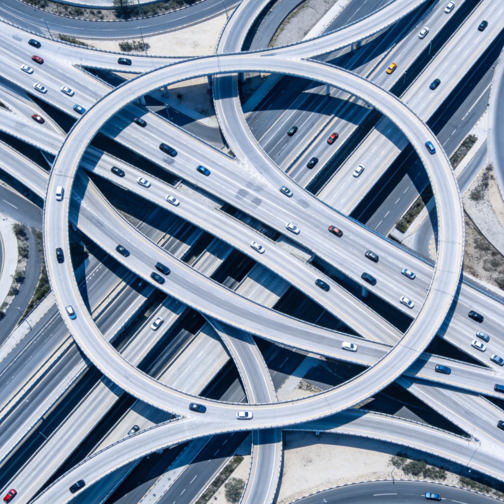 An aerial view of a clean, modern city at daytime, focusing on the intricate layout of roads, highways, and intersections that connect different areas. Some tiny cars are visible on the roads. The perspective emphasizes the "network" pattern. No text.