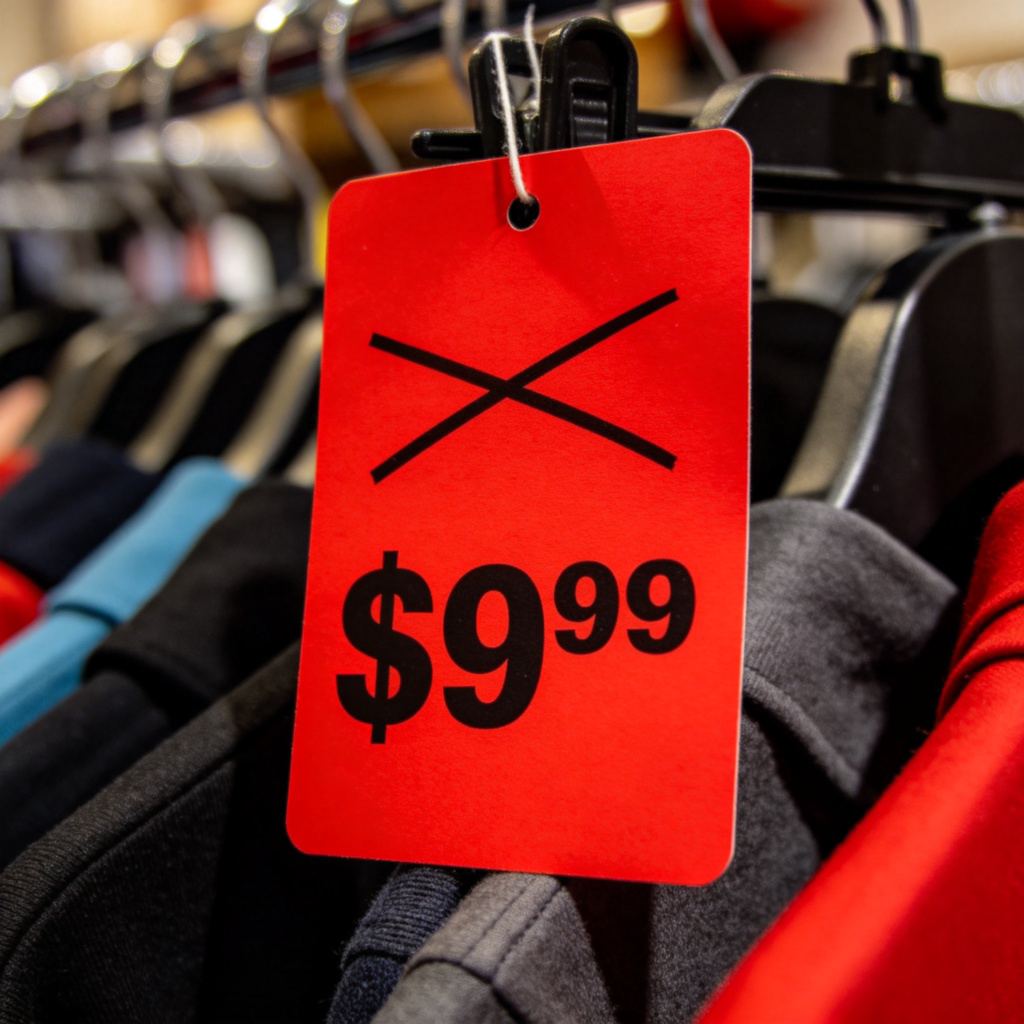 A close-up of a bright red price tag on a piece of clothing in a store. The original higher price is clearly crossed out in black, and a new, lower price is written boldly below it. The background is slightly blurred fabric racks, emphasizing the tag. No text other than the prices.